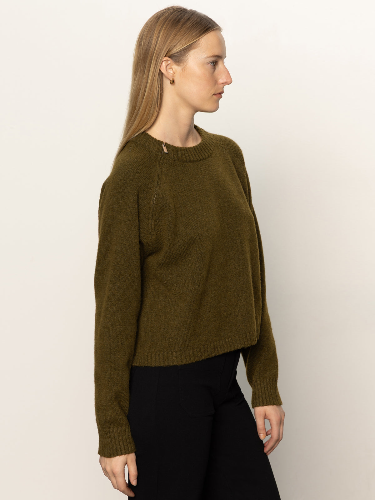 A woman with long blonde hair wears the Sanctuary Clothing "zip it sweater dirty martini" in dark olive green, paired with black pants, standing in profile against a plain light background.