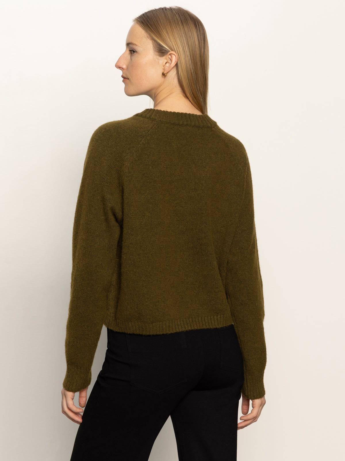 A woman with straight, light brown hair stands facing away against a plain background, wearing Sanctuary Clothing's zip it sweater in dirty martini (olive green), paired with black pants.
