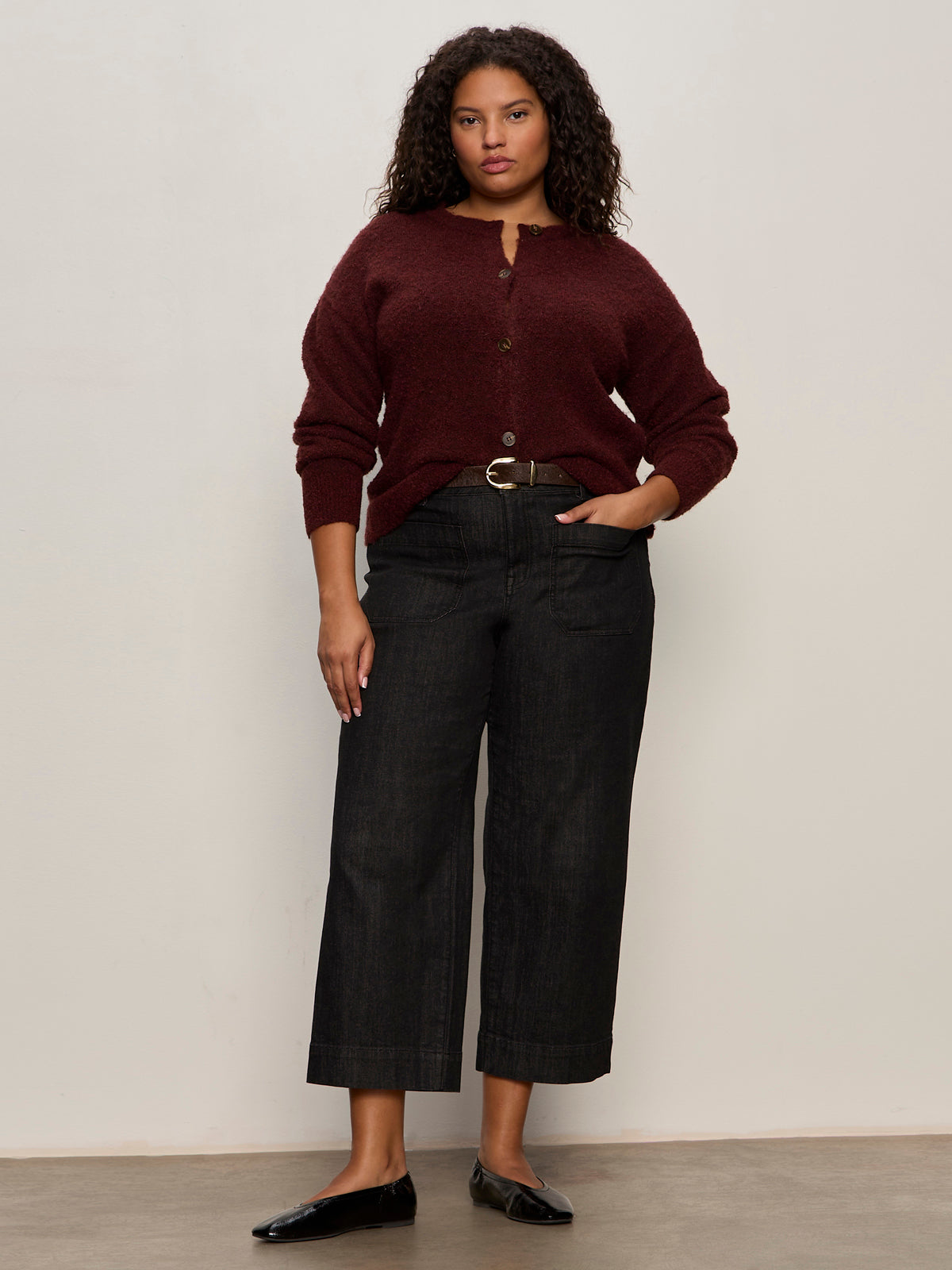A woman with curly hair wears the Sanctuary Clothing boucle cardi in dark crimson (extended sizing), paired with dark wide-leg jeans, a black belt, and black flats, standing against a plain light-colored wall.