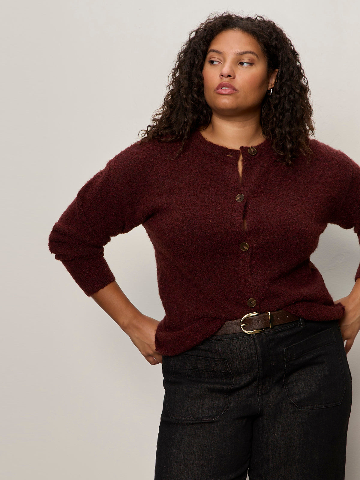A woman with curly hair models the Sanctuary Clothing boucle cardi in dark crimson (extended sizing), paired with dark high-waisted jeans, standing against a plain light background with one hand on her hip and a neutral expression.