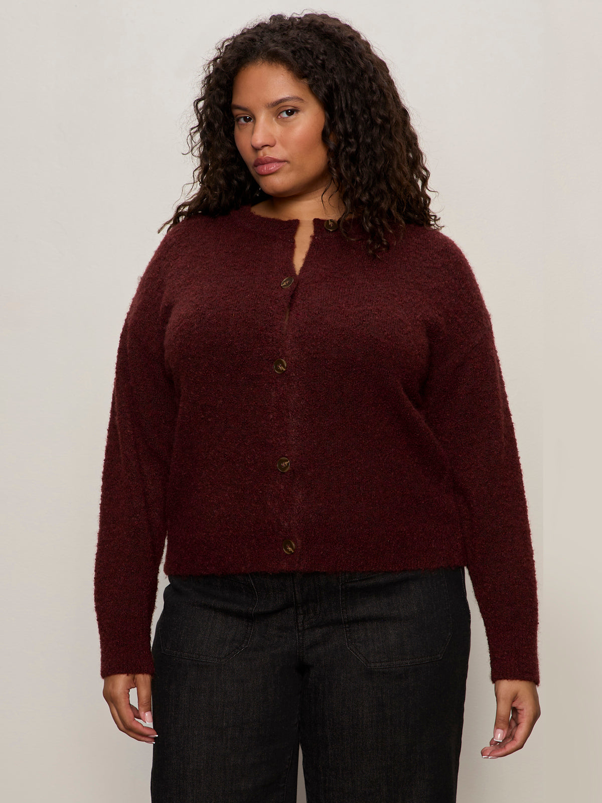A woman with curly hair wears Sanctuary Clothing's boucle cardi in dark crimson (extended sizing) and black pants, standing against a plain, light-colored background.