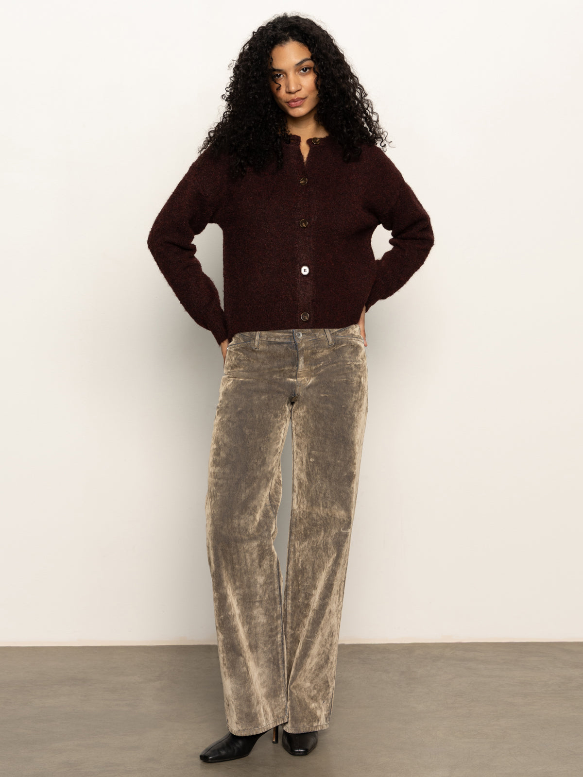 A person with long curly hair stands against a plain background, wearing Sanctuary Clothing„¢s boucle cardi in dark crimson, beige velvety wide-leg pants, and black shoes, with hands resting on their hips.