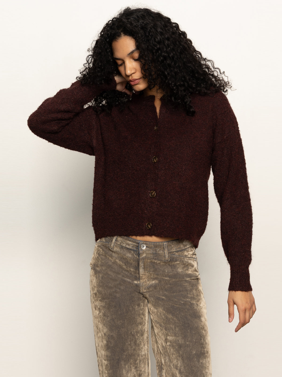A person with long curly hair wears the Sanctuary Clothing boucle cardi in dark crimson and beige corduroy pants, standing against a light background, looking down with one hand touching their head.
