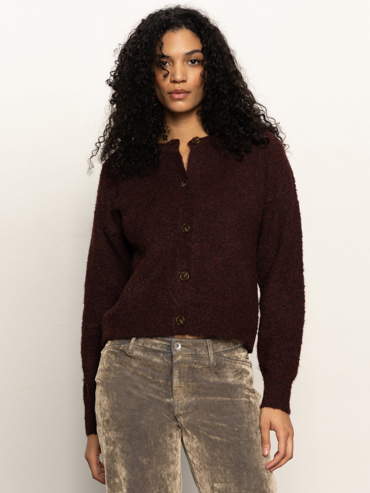 A woman with curly dark hair wears the Sanctuary Clothing boucle cardi in dark crimson and light brown corduroy pants, standing against a plain light background.
