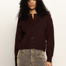 A woman with curly dark hair wears the Sanctuary Clothing boucle cardi in dark crimson and light brown corduroy pants, standing against a plain light background.