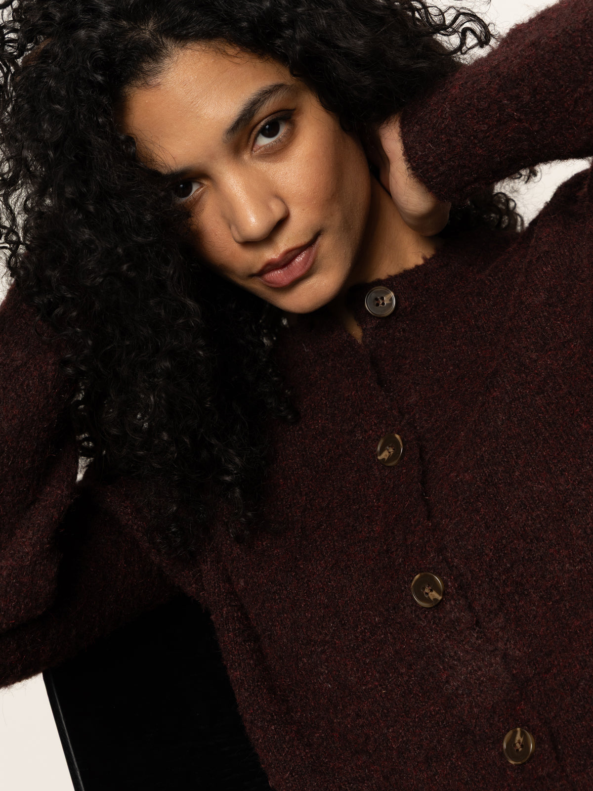 A woman with curly dark hair poses with her hands near her head, modeling Sanctuary Clothing„¢s boucle cardi in dark crimson and looking at the camera against a plain background.