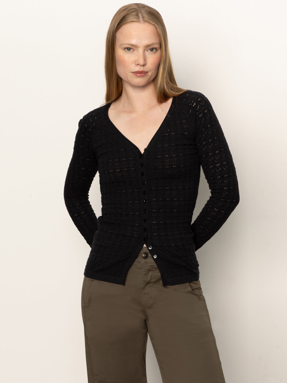 A woman with straight, light brown hair models the Sanctuary Clothing fitted raglan cardi in black, paired with olive green pants, against a plain light background.