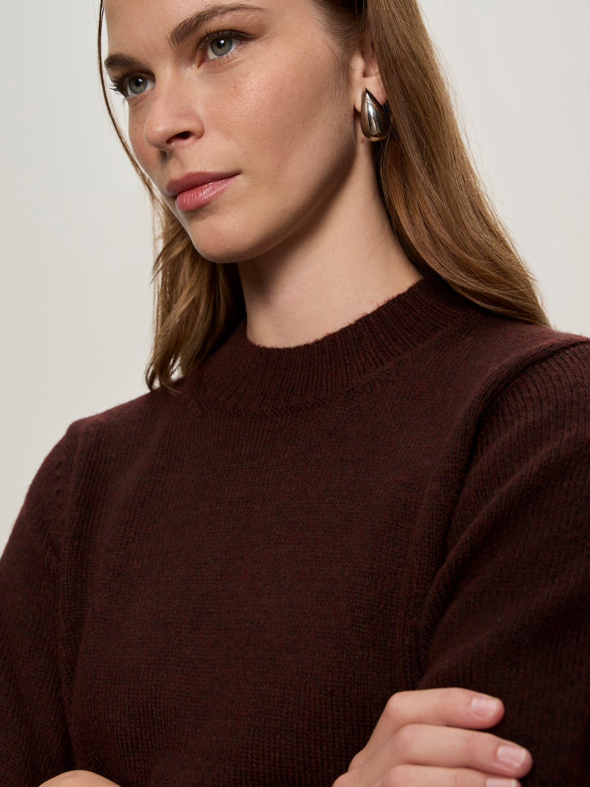 A woman with light brown hair and silver hoop earrings stands with her arms crossed, wearing the Sanctuary Clothing perfect sweater tee in dark crimson, gazing slightly to the side with a neutral expression.