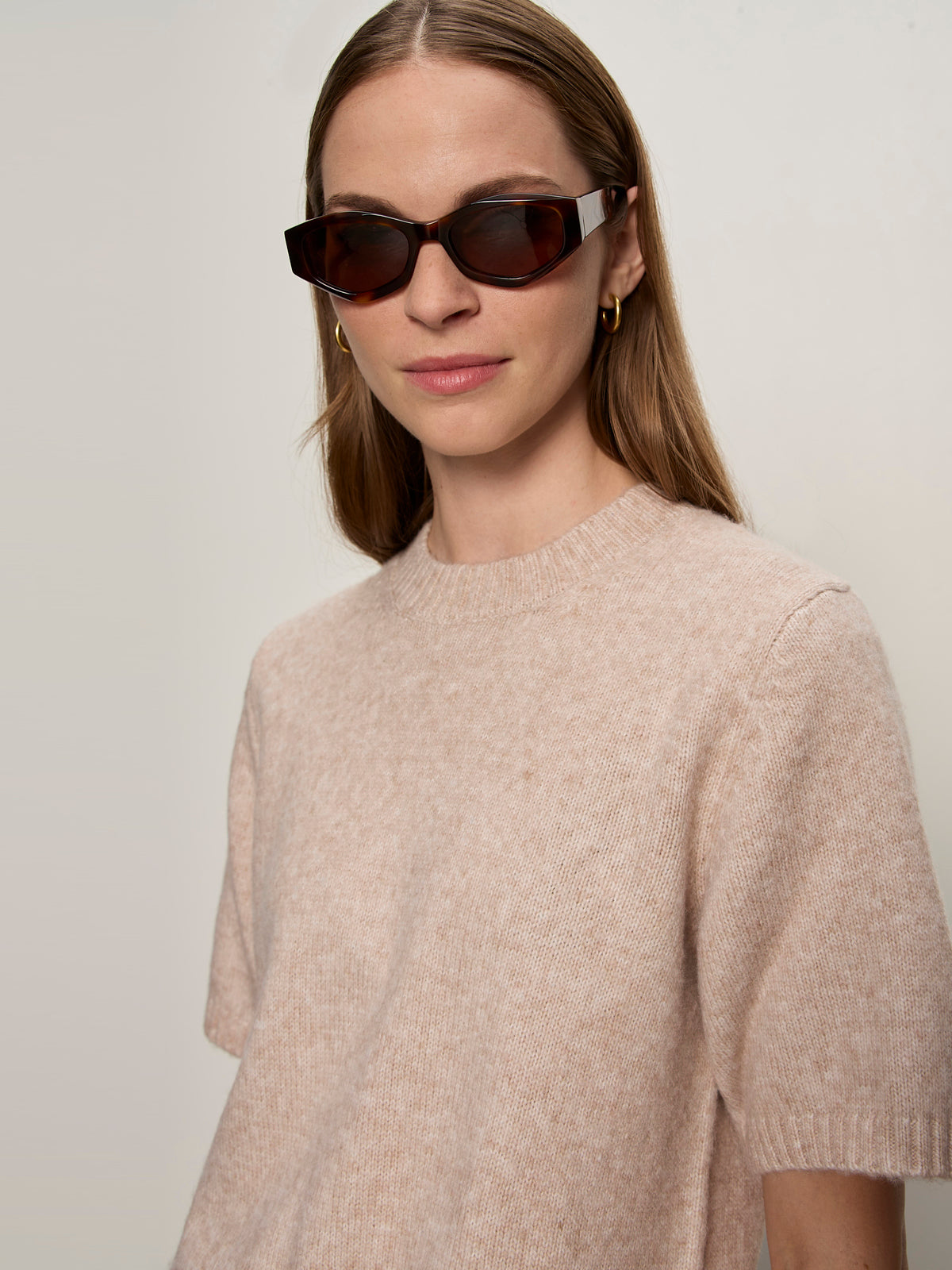 A woman with straight, light brown hair wears dark rectangular sunglasses, gold hoop earrings, and the Sanctuary Clothing perfect sweater tee in fawn, standing against a plain light background.