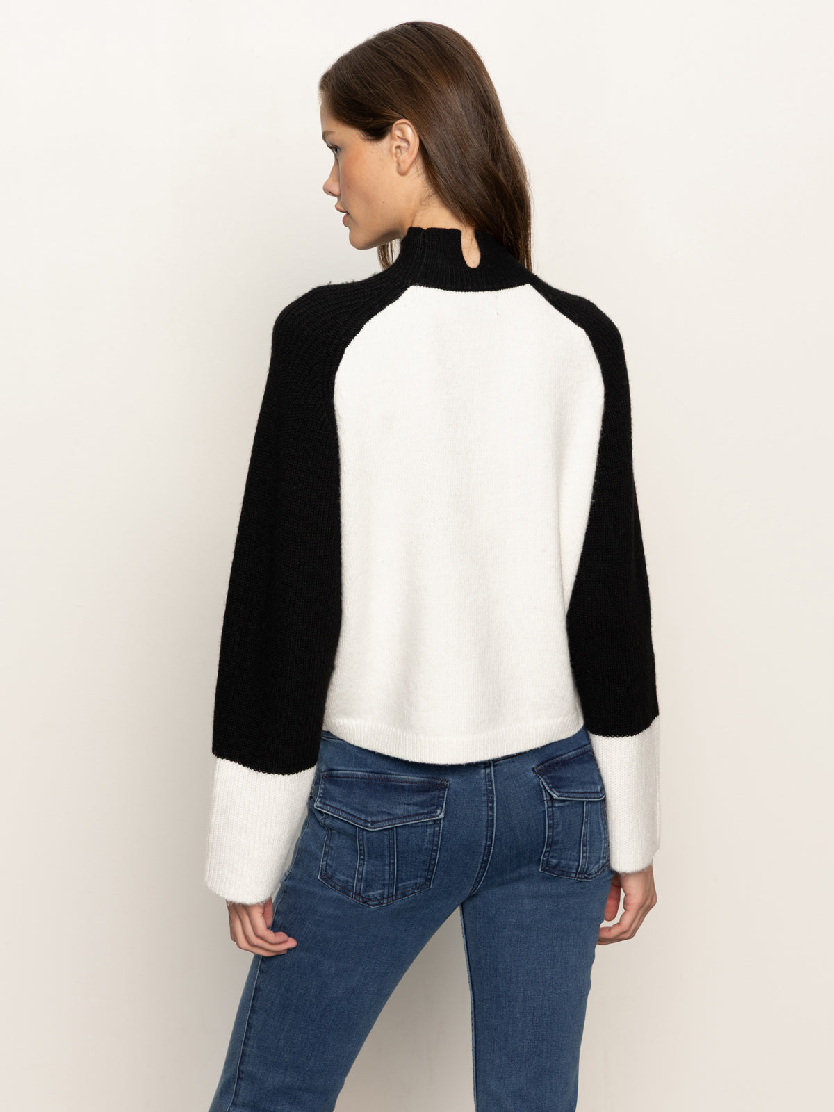 A woman with long brown hair, seen from behind, wears the Sanctuary Clothing downtown mock neck sweater in black chalk with wide sleeves and blue jeans, standing against a plain light background.
