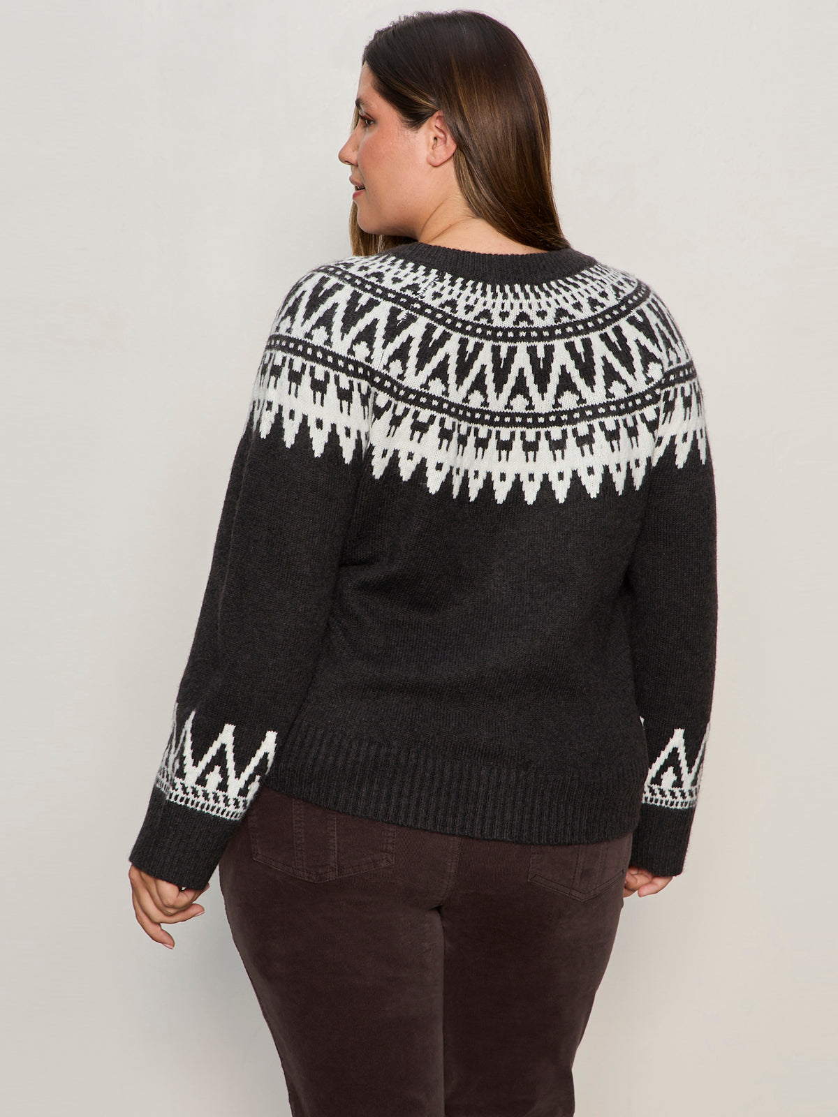 A woman with long brown hair, dressed in Sanctuary Clothingžs fairisle crewneck cardi ink multi extended sizing and dark brown pants, stands facing away and looks over her left shoulder.