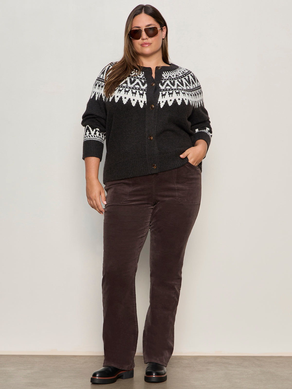 A woman wearing Sanctuary Clothing's fairisle crewneck cardi in ink multi (extended sizing), sunglasses, brown corduroy pants, and black shoes stands against a plain light wall with one hand in her pocket and a relaxed posture.