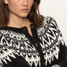 A woman with wavy brown hair wears the Sanctuary Clothing fairisle crewneck cardi in ink multi, featuring a black and white pattern and wooden buttons, as she looks seriously at the camera against a plain background.