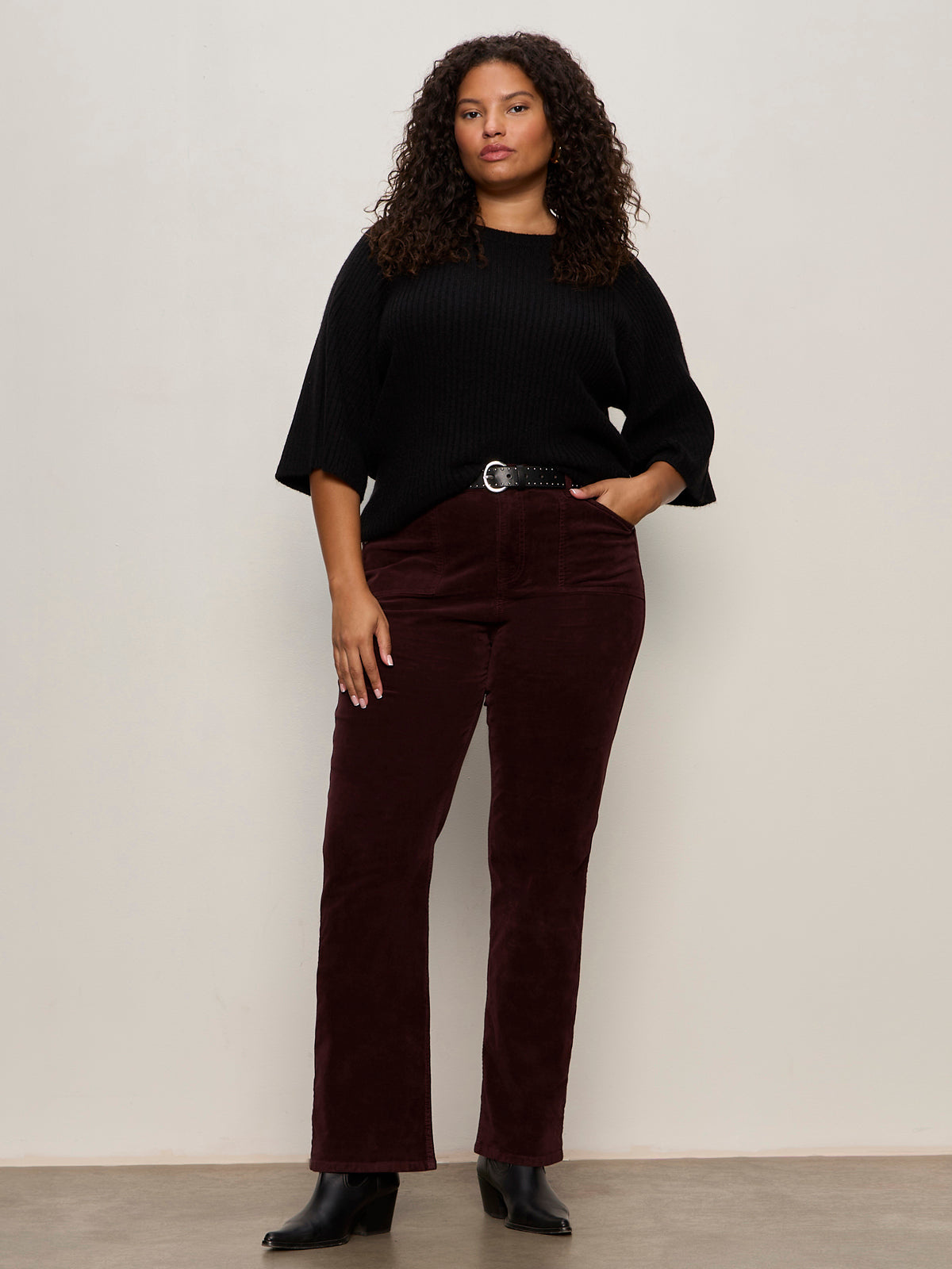 A woman with curly hair wears a Sanctuary Clothing ribbed sweater tee in black (extended sizing), dark burgundy corduroy pants, a black belt, and black ankle boots while standing against a plain light-colored wall.