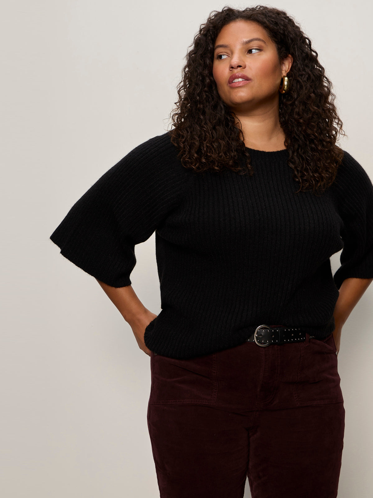 A woman with curly hair wears the Sanctuary Clothing ribbed sweater tee in black (extended sizing) with dark maroon pants and a black belt, standing against a plain light background with her hands behind her back.