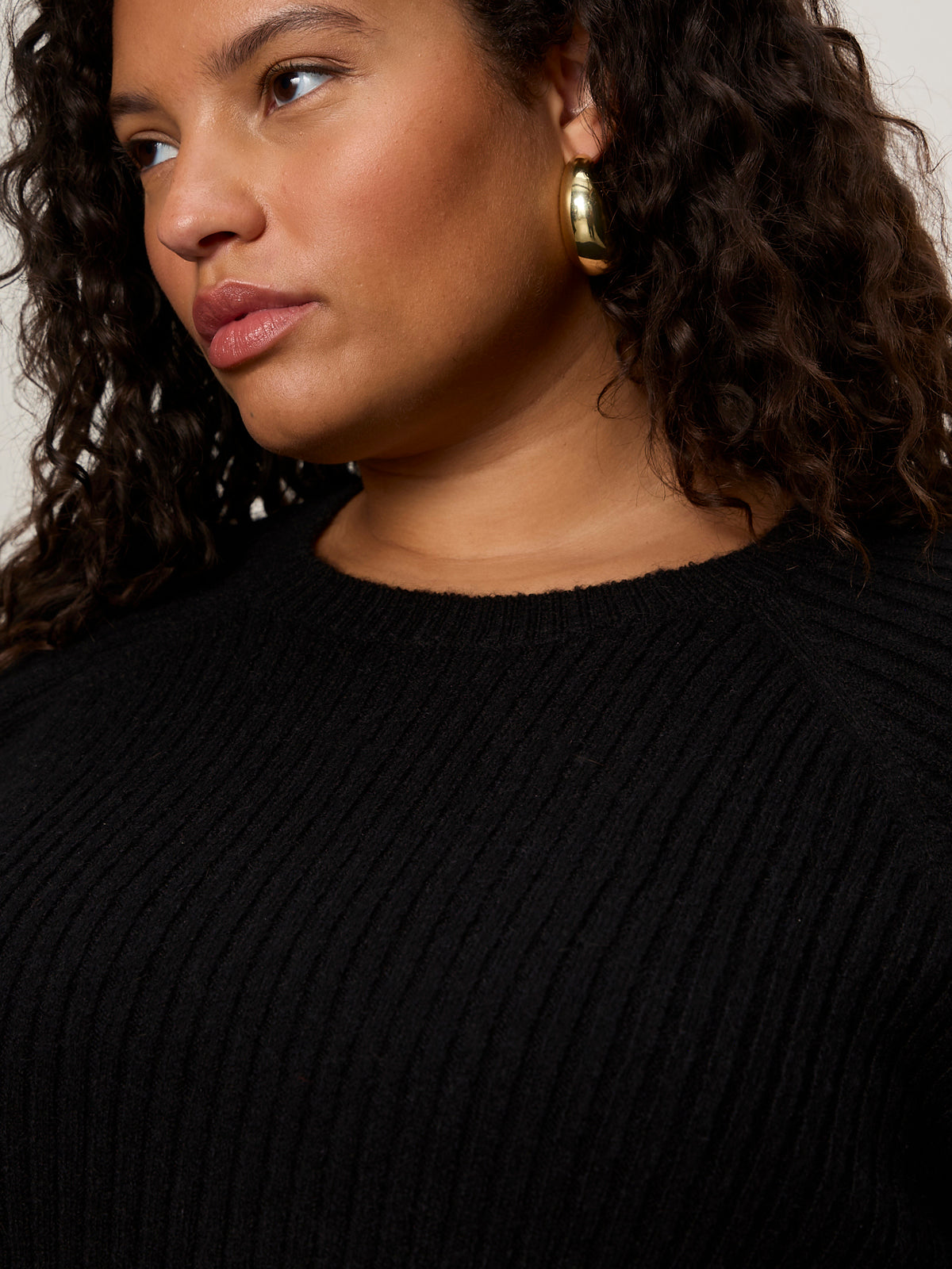 A person with curly hair wears the Sanctuary Clothing ribbed sweater tee in black (extended sizing), paired with large gold hoop earrings, looking slightly to the side in a closely cropped upper torso and face image.