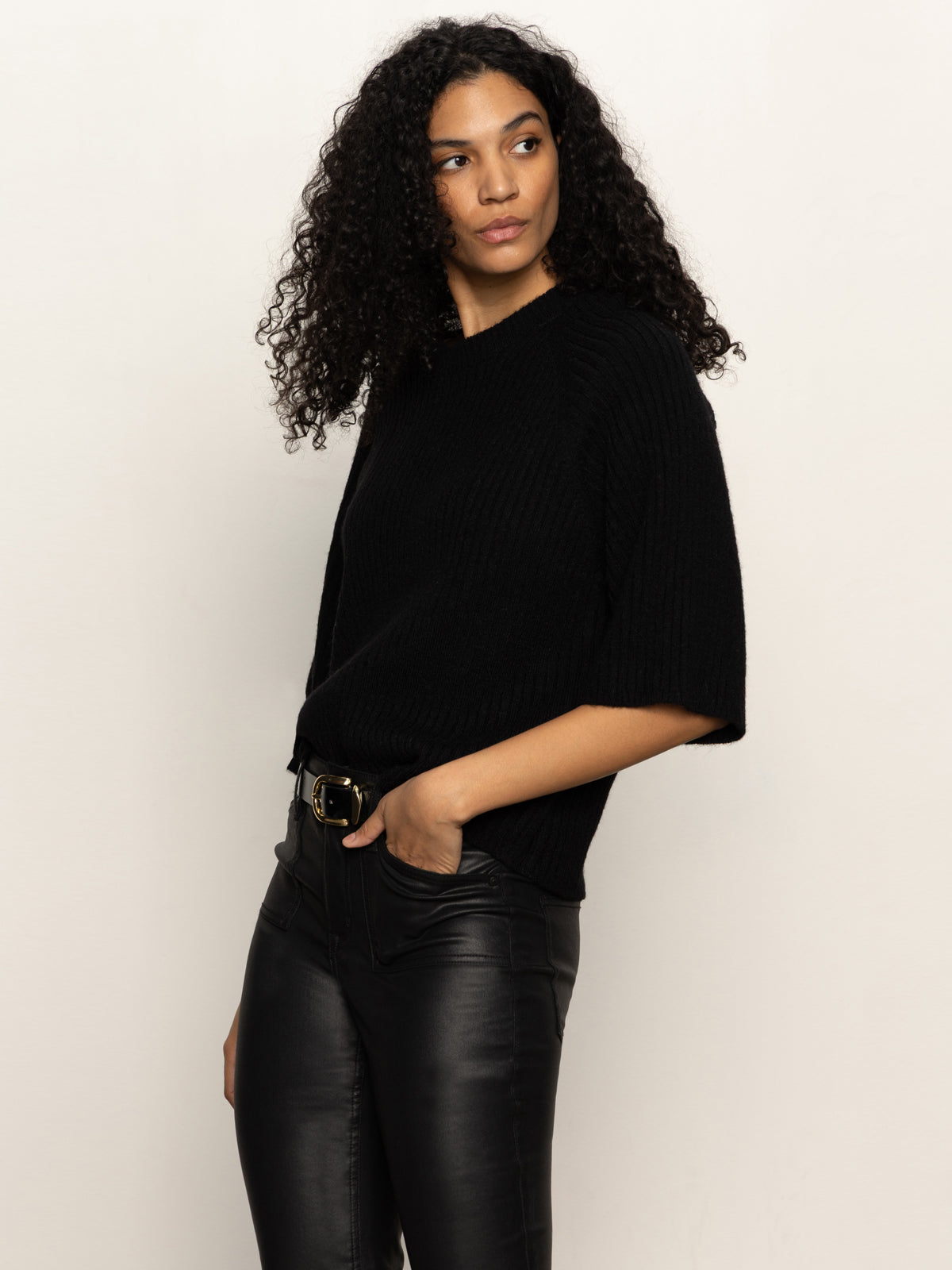 A woman with curly hair models the Sanctuary Clothing ribbed sweater tee in black, paired with black leather pants and posing with one hand in her pocket against a plain, light background.