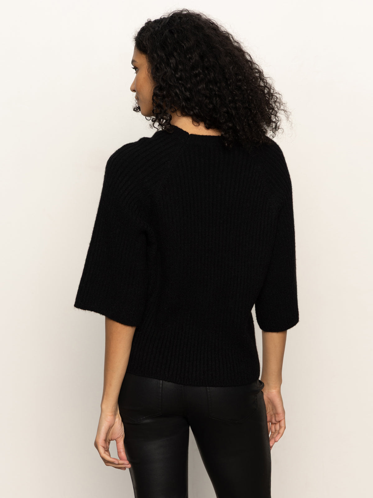 A woman with curly hair faces away from the camera, wearing the Sanctuary Clothing ribbed sweater tee in black against a plain light background.
