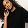 A woman with curly hair, wearing the Sanctuary Clothing ribbed sweater tee in black, poses against a light background, tilting her head and gently touching her face with one hand.