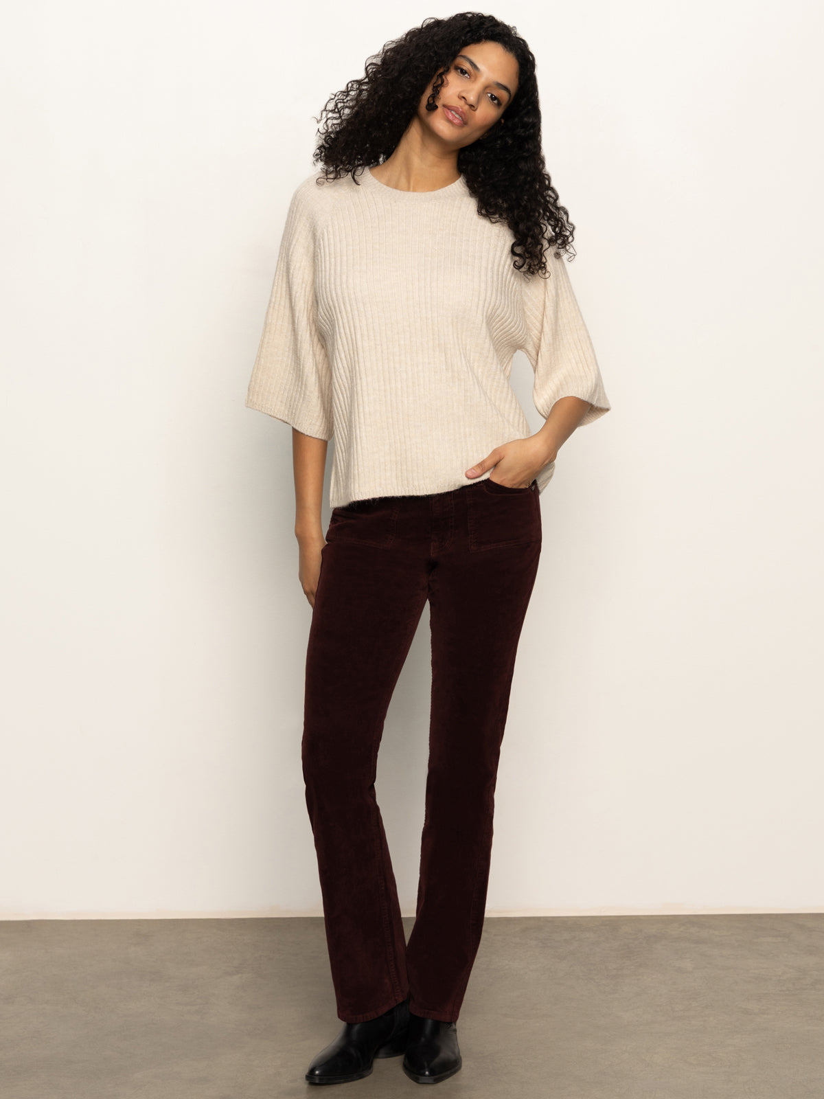 A woman with curly hair wears the ribbed sweater tee mother of pearl by Sanctuary Clothing, paired with dark red corduroy pants and black shoes. She stands against a plain light background, hand in her pocket and looking relaxed.
