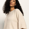 A woman with long, curly dark hair wears the Sanctuary Clothing ribbed sweater tee in mother of pearl, featuring elbow-length sleeves. She stands against a plain, light background and gazes slightly downward with a neutral expression.