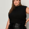 A woman with long brown hair models the Sanctuary Clothing city sweater shell black (extended sizing), paired with a black leather skirt. She stands against a light background, looking confidently at the camera with one hand on her hip.