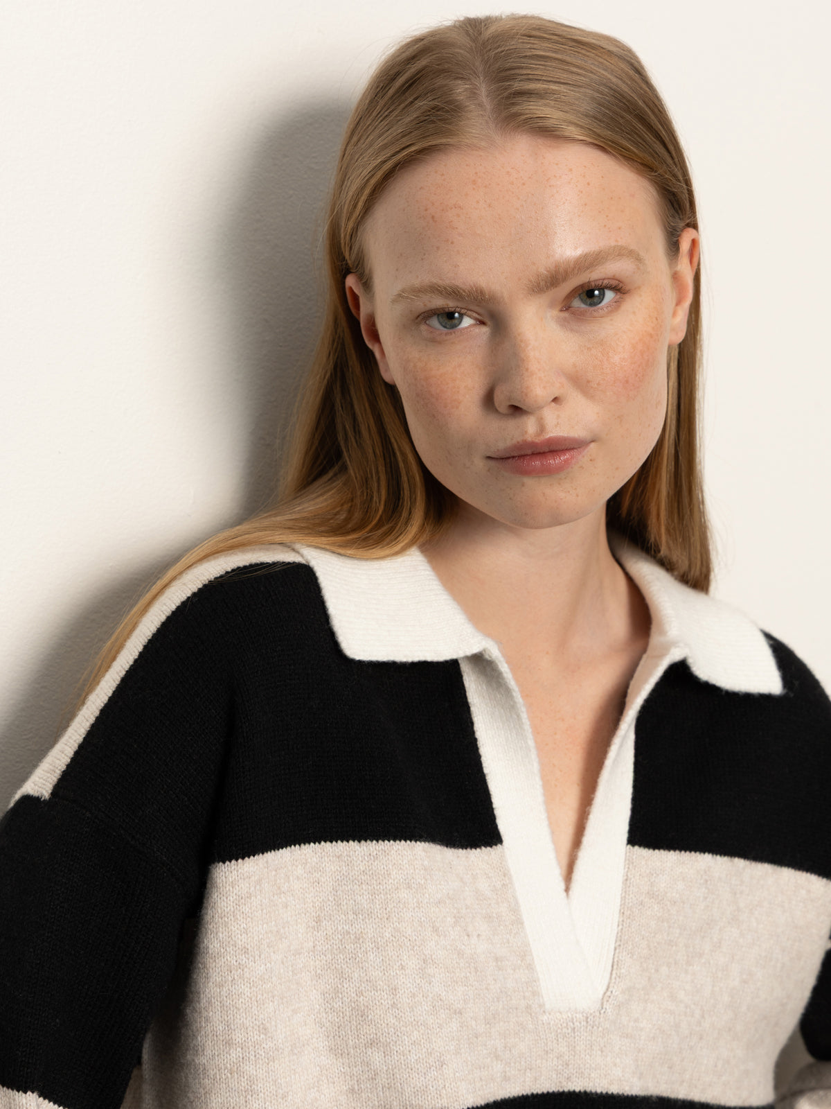 A woman with long blonde hair and fair skin wears the Sanctuary Clothing polo striped sweater dress in heather pearl with black, standing against a light-colored wall and looking directly at the camera with a neutral expression.