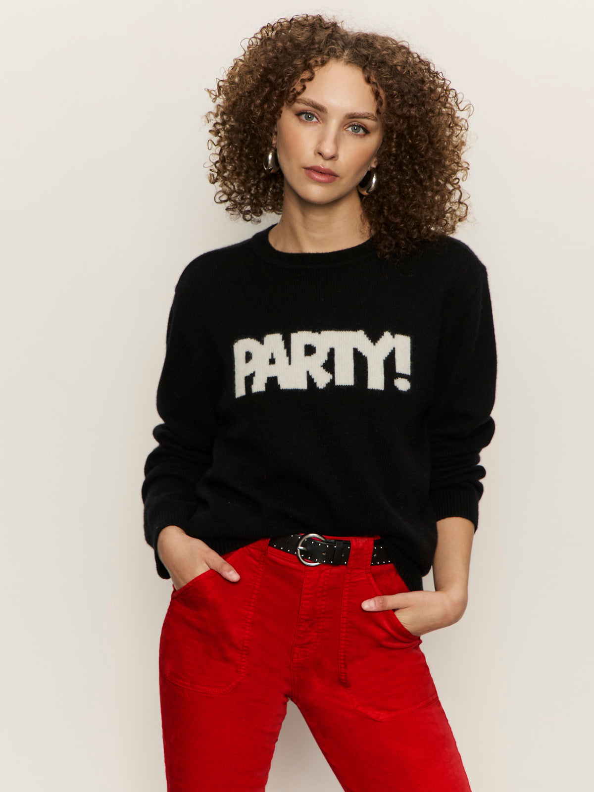 A woman with curly hair wears the Sanctuary Clothing "life of the party" cashmere sweater in black with chalk lettering, paired with bright red pants, standing against a plain light background with her hands in her pockets.