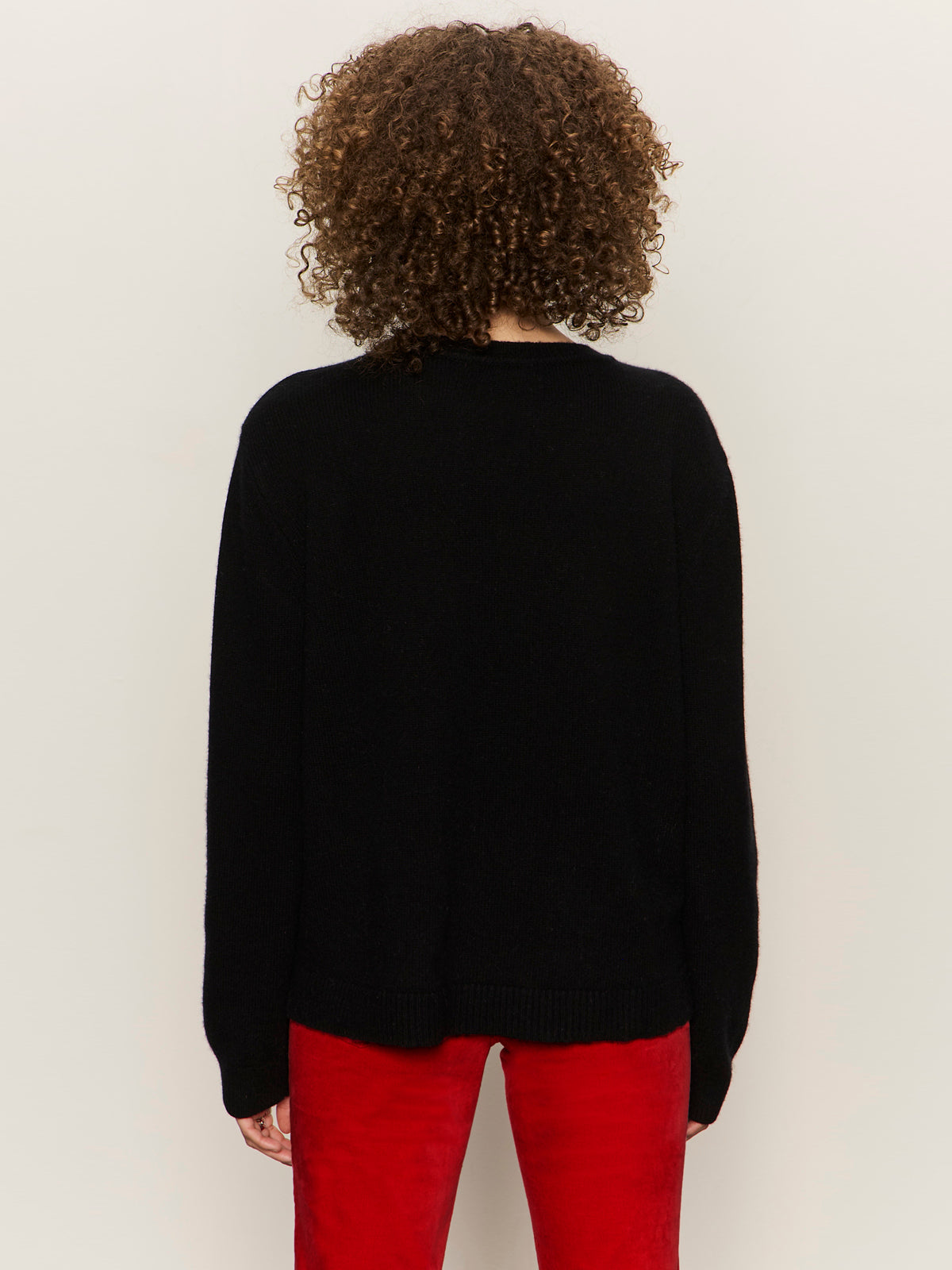 A person with curly brown hair is seen from behind wearing the Sanctuary Clothing life of the party cashmere sweater in black with chalk, paired with red pants, against a plain, light background.