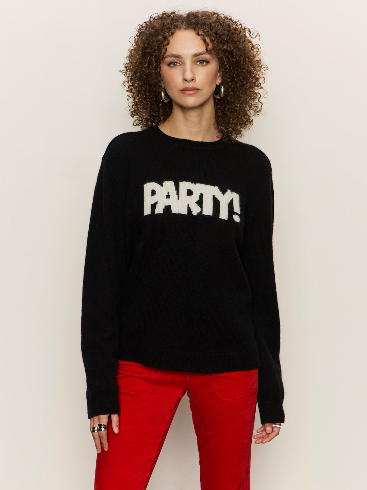 A woman with curly hair wears the Sanctuary Clothing "life of the party" black cashmere sweater with chalk lettering, paired with bright red pants. She stands against a plain light background, looking directly at the camera.