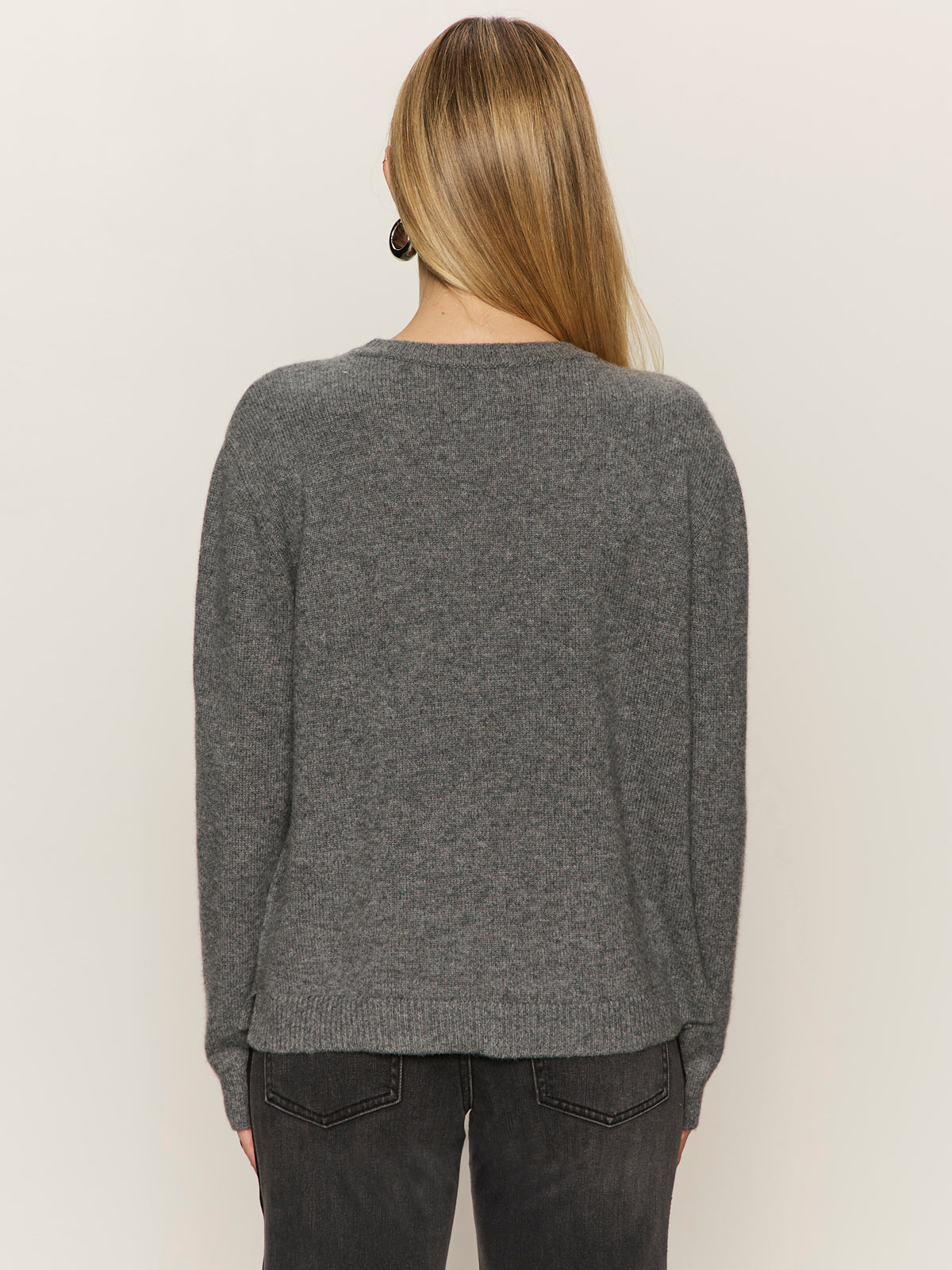 A person with long, straight blonde hair wears the Sanctuary Clothing life of the party cashmere sweater in heather charcoal with chalk, paired with dark jeans, standing with their back to the camera against a plain light background.
