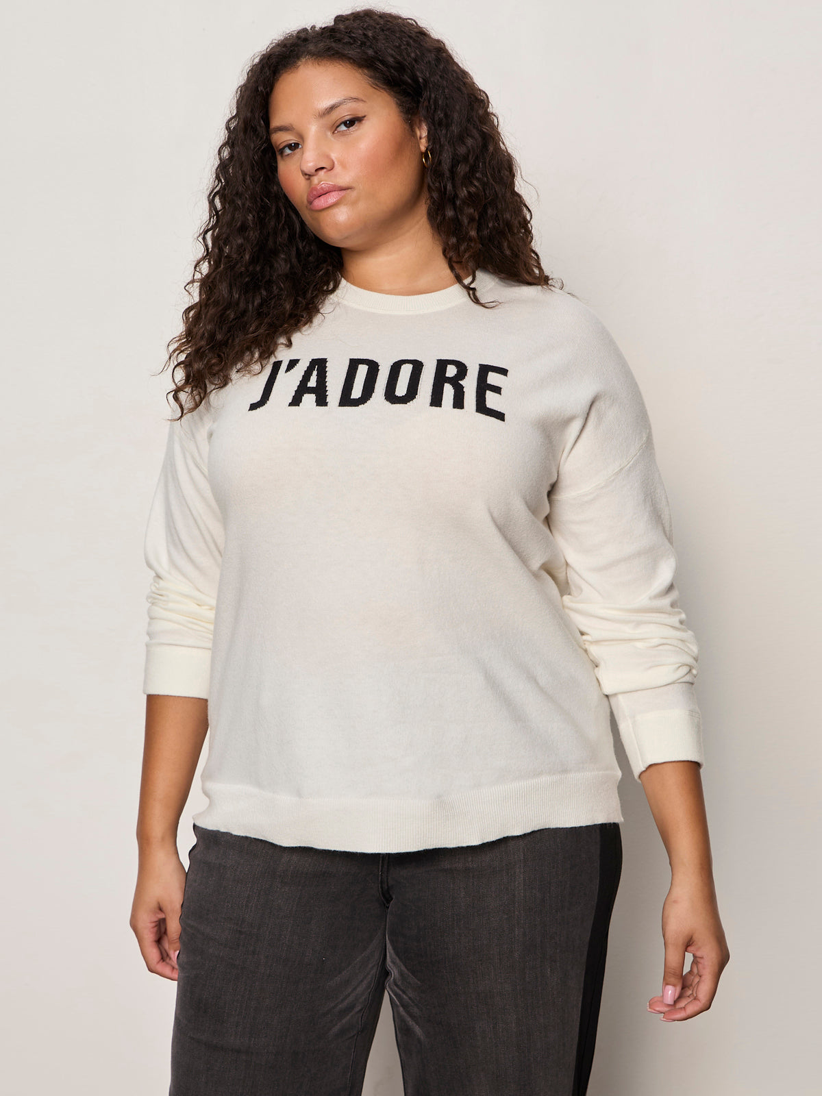 A woman with curly hair wears a cream-colored sweater with the word "J'ADORE" in bold black letters, paired with dark pants, standing against a plain light background.