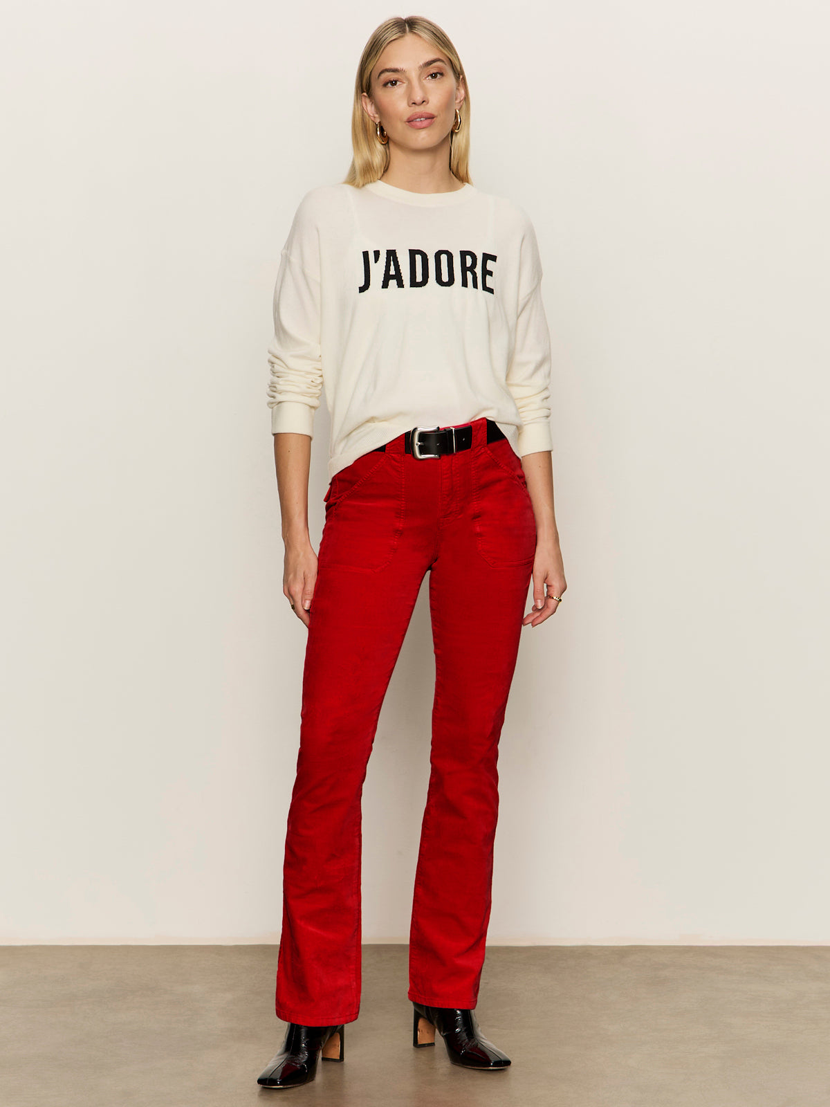 A woman with blonde hair and a relaxed expression wears the Sanctuary Clothing "24 7 popover chalk with black" sweater against a plain background, paired with red pants, a black belt, and black boots.