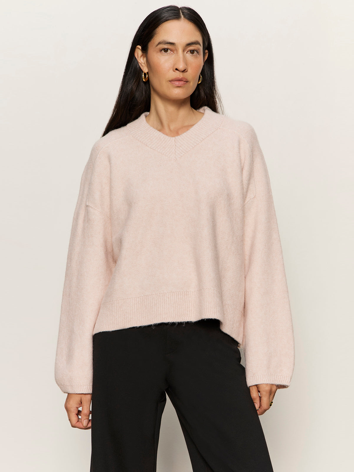 A woman with long dark hair wears the Sanctuary Clothing dreamy v neck sweater in heather rose and black pants, standing against a light background with a neutral expression, facing forward.