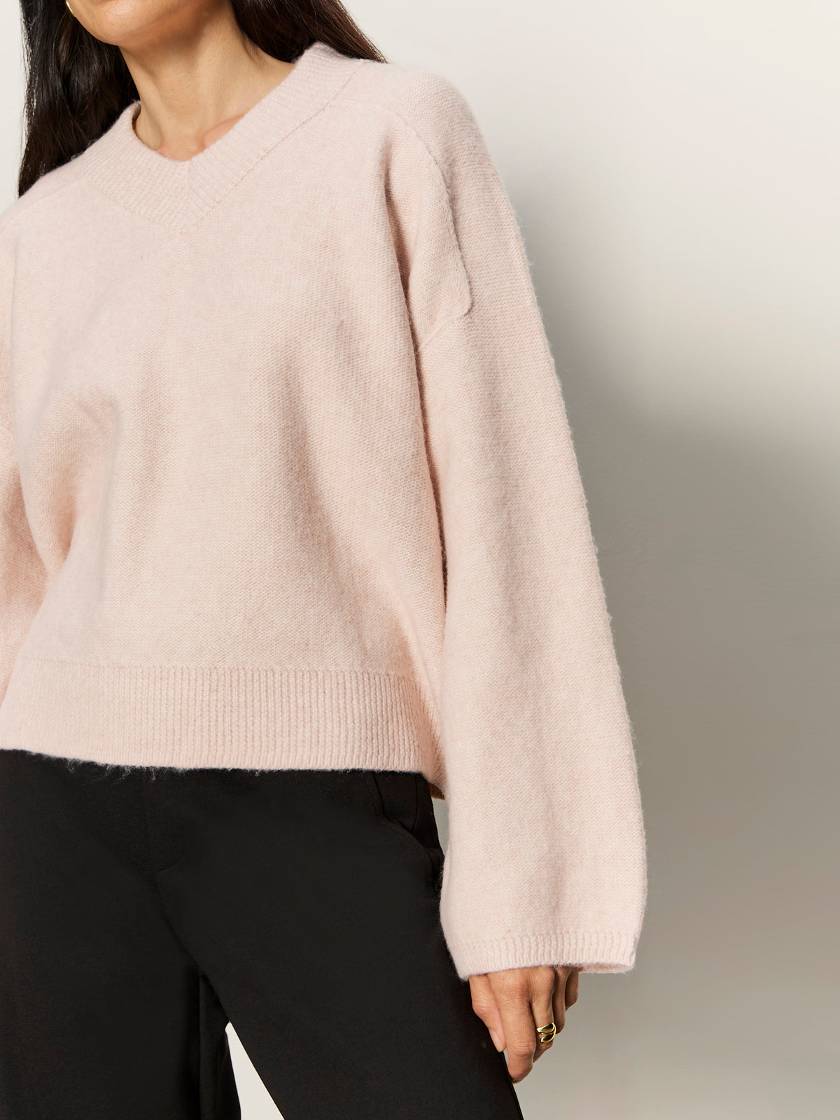 A person wearing the Sanctuary Clothing dreamy v neck sweater in heather rose with black pants stands against a plain light background, their face partially out of frame.