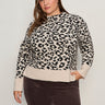 A woman wears the Sanctuary Clothing soft touch popover spots extended sizing sweater in a beige and black leopard print with long sleeves, paired with dark brown pants, standing against a plain light wall and smiling slightly at the camera.
