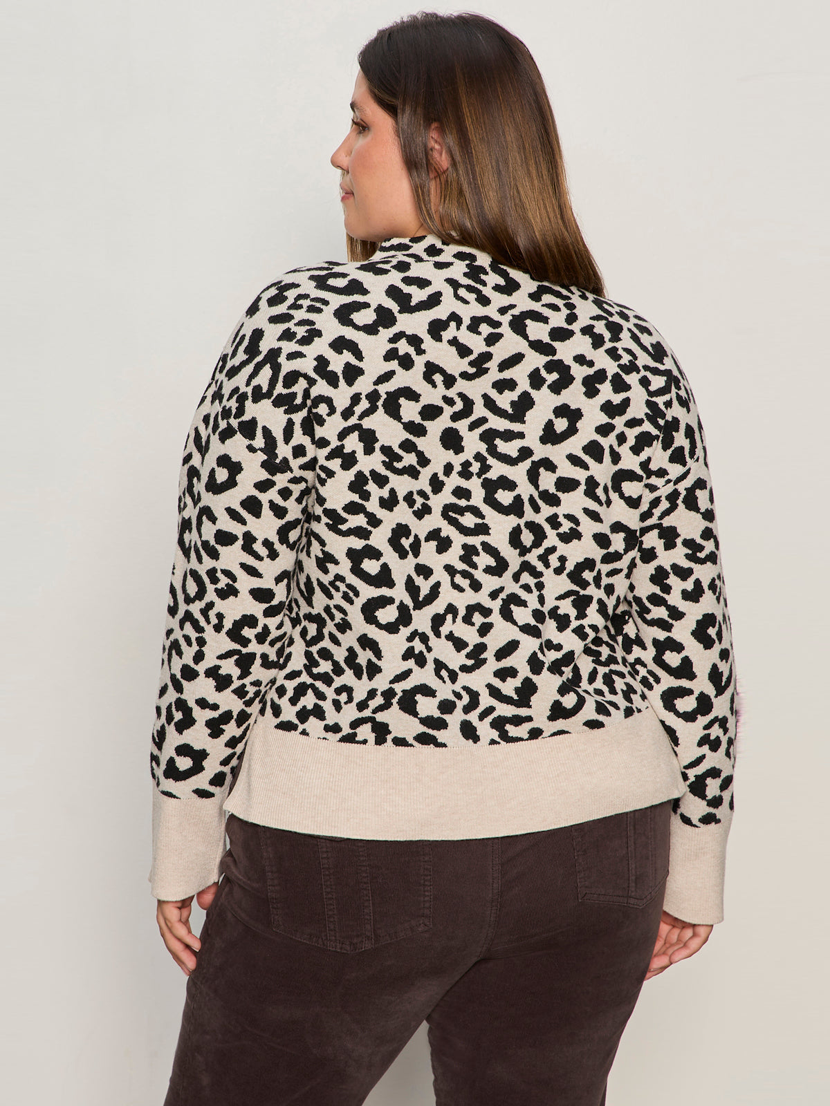 A woman with long brown hair is seen from the back wearing the Sanctuary Clothing soft touch popover spots sweater in beige with a black leopard print and dark brown pants, standing against a plain light-colored wall.