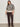 A woman with long brown hair and a relaxed, confident expression wears the Sanctuary Clothing soft touch popover spots extended sizing sweater against a plain light wall, styled with dark brown corduroy pants and black shoes.