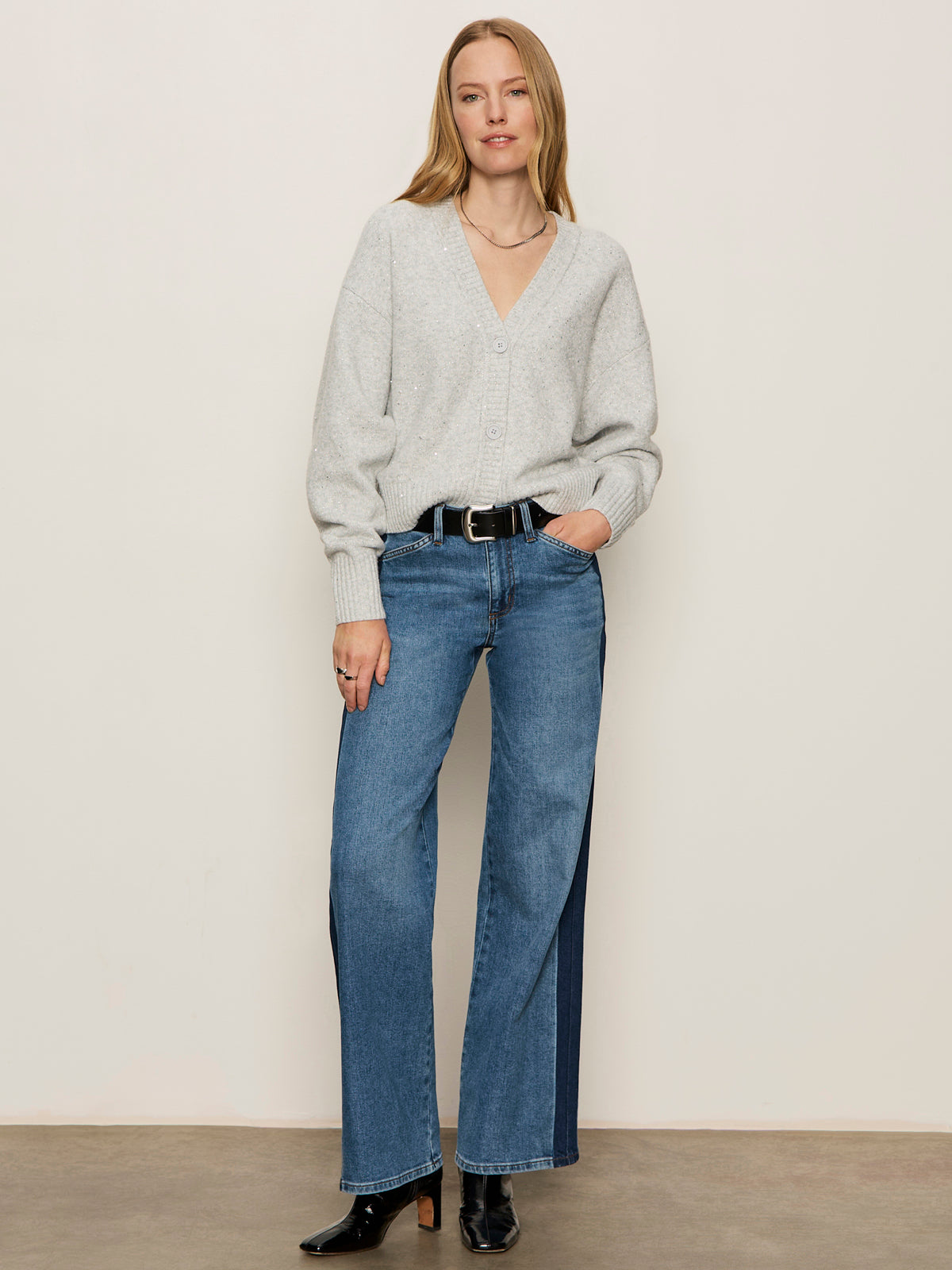 A woman stands against a plain wall, wearing the Sanctuary Clothing shimmer cardi in heather grey sparkle, paired with blue high-waisted jeans, a black belt, and black ankle boots. Her straight blonde hair frames her relaxed, confident expression.