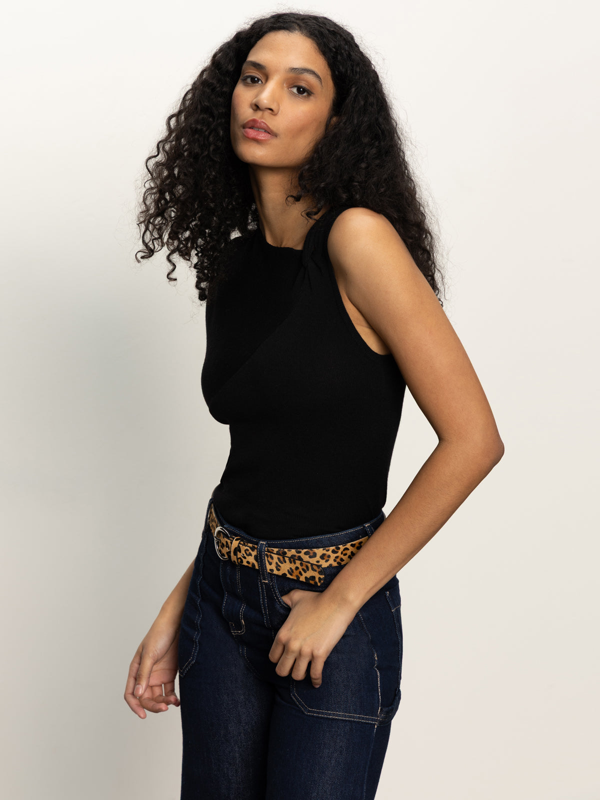 A woman with long curly hair wears a sleeveless black top, dark jeans, and the Sanctuary Clothing riot leopard belt leopard, standing against a plain light background.