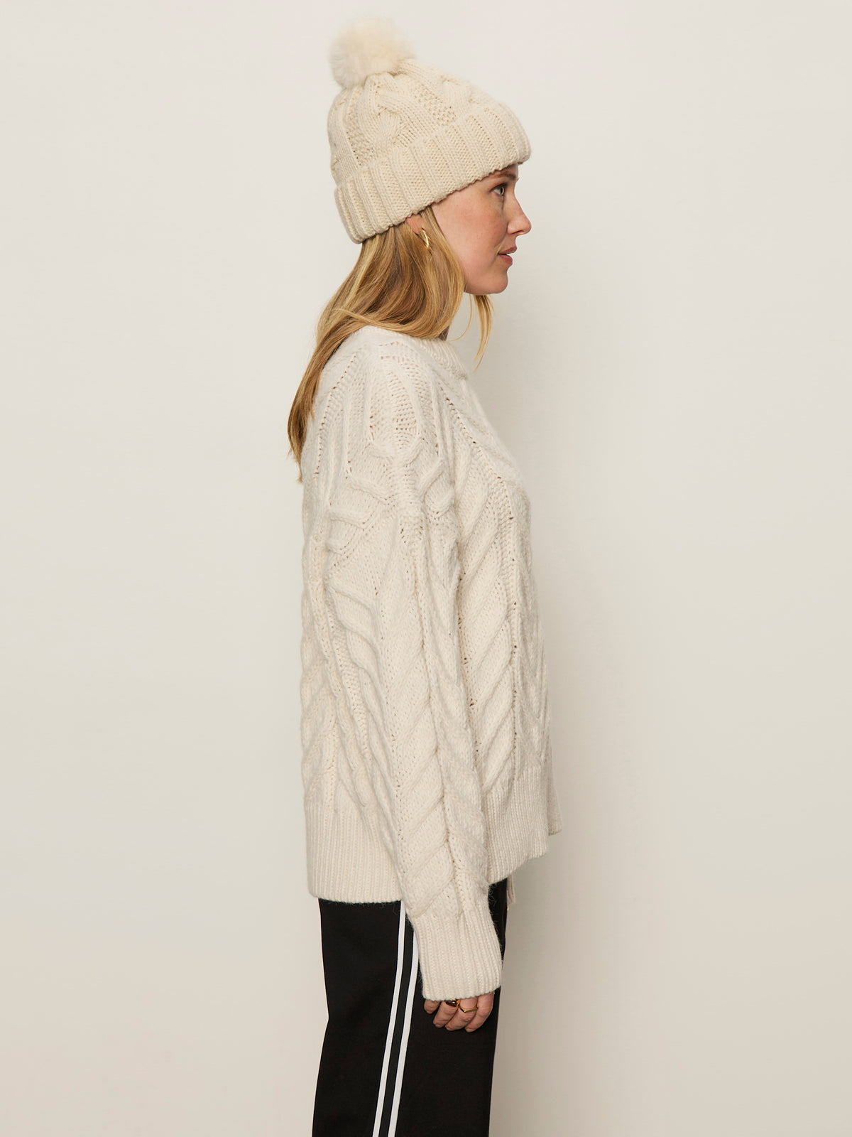 A woman with long blonde hair wears the Sanctuary Clothing snow bunny statement sweater in chalk—a cream cable-knit, paired with a matching pom-pom beanie and black pants with white stripes, against a light background.