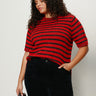 A woman with curly hair wears the Sanctuary Clothing short sleeve shine sweater tee in cherry lacquer shine (extended sizing) and black pants. She stands against a light background, one hand in her pocket, holding a black studded bag.