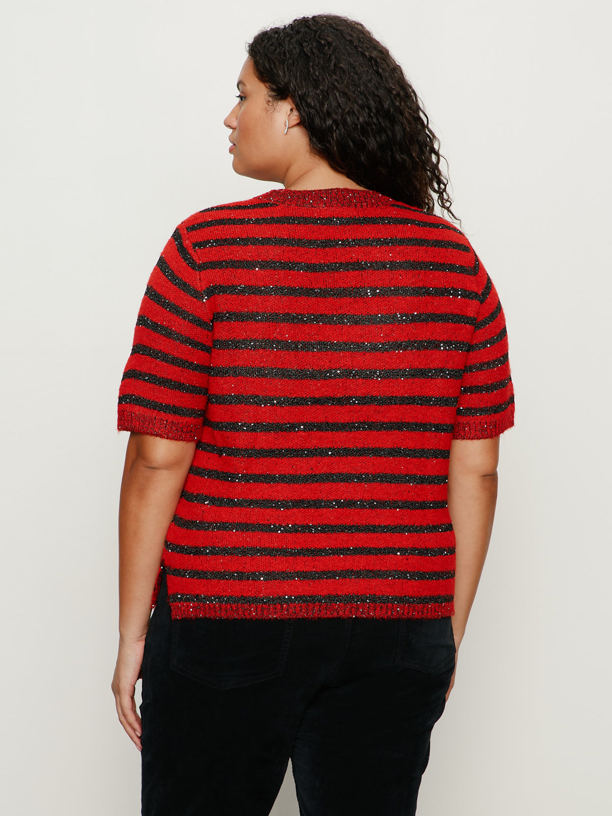 A person with curly hair is seen from behind, wearing Sanctuary Clothing's short sleeve shine sweater tee in cherry lacquer shine (extended sizing) and black pants, standing against a plain light background.