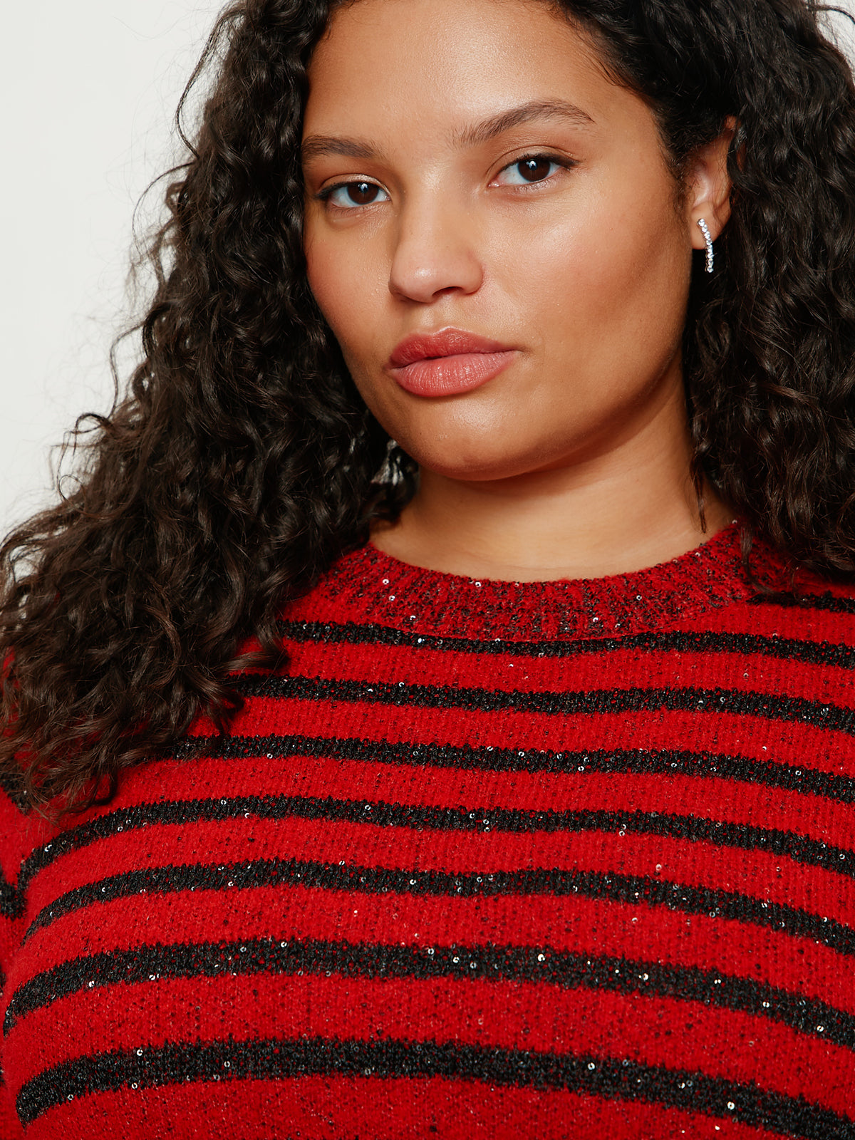A person with curly dark hair wears the Sanctuary Clothing short sleeve shine sweater tee in cherry lacquer shine extended sizing, featuring black horizontal stripes and subtle sparkles, looking directly at the camera with a neutral expression.