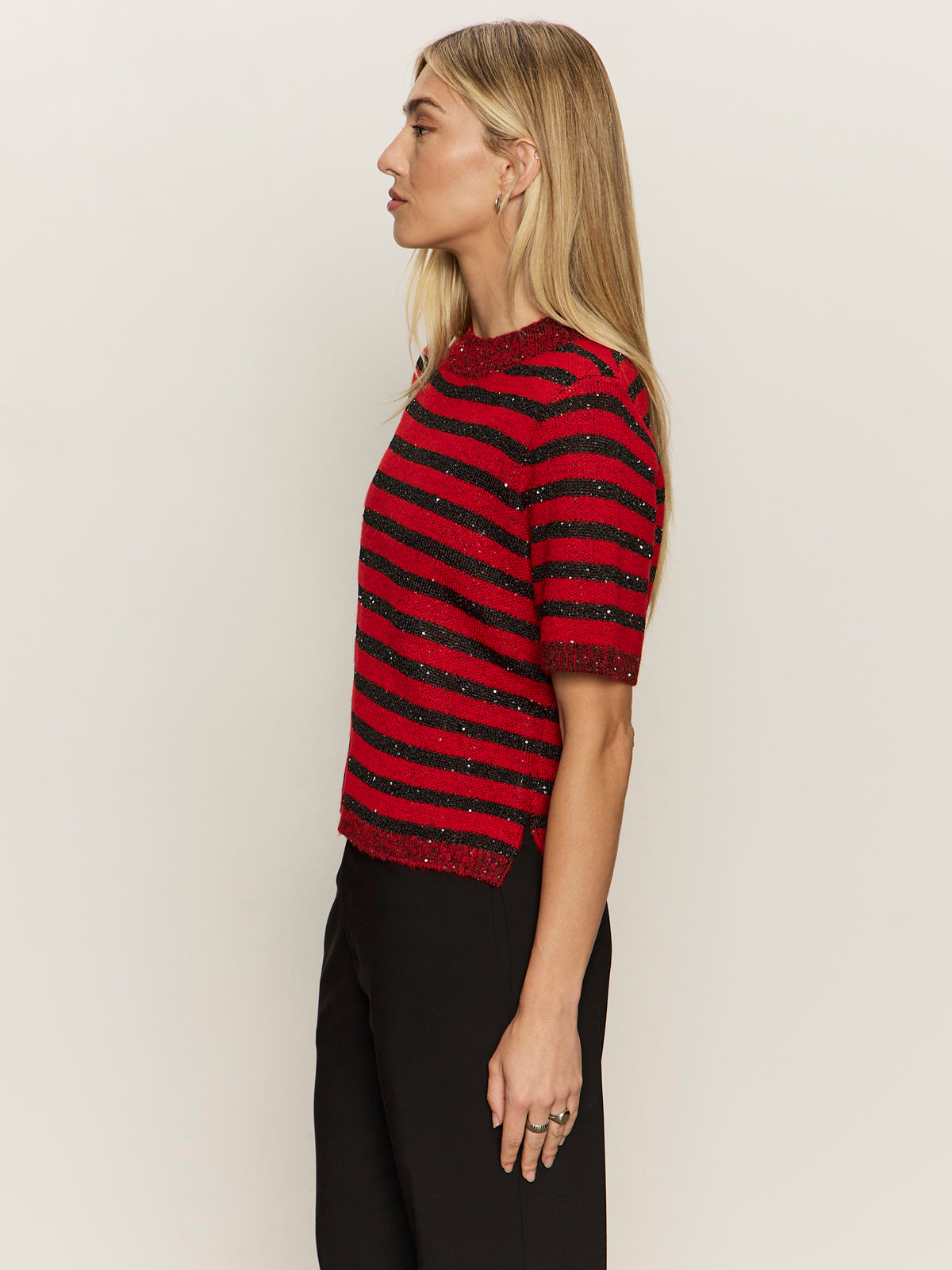 A woman with long blonde hair stands in profile, wearing the Sanctuary Clothing short sleeve shine sweater tee in cherry lacquer shine and black pants against a plain light background.