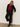 A person with long blonde hair leans against a beige wall, wearing Sanctuary Clothing's short sleeve shine sweater tee in cherry lacquer shine, paired with black pants featuring ankle button details and black heeled ankle boots.