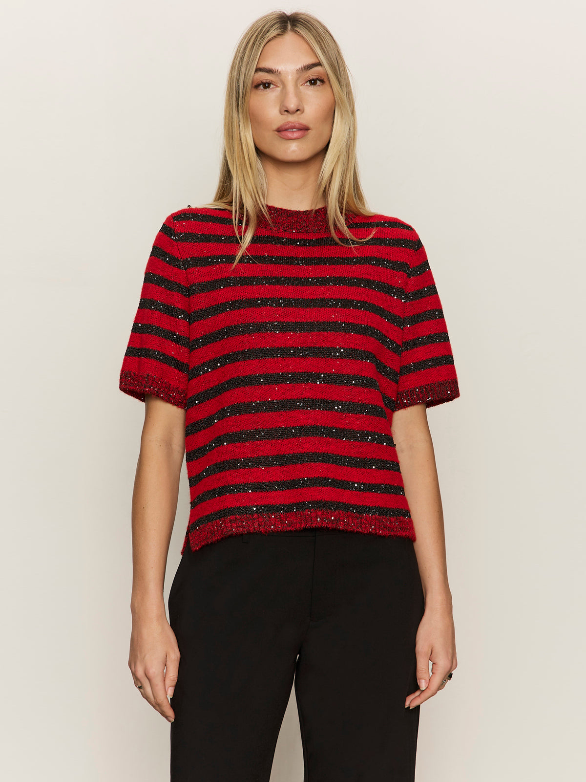 A woman with straight blonde hair wears the Sanctuary Clothing short sleeve shine sweater tee in cherry lacquer shine, featuring red and black horizontal stripes with subtle sparkle, paired with black pants against a light background.