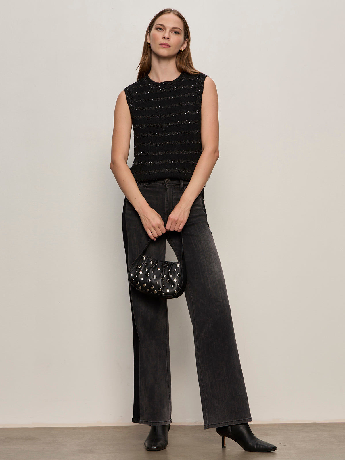A woman stands against a plain light-colored wall wearing the Sanctuary Clothing shine sweater shell black with black shine, wide-leg dark jeans, black heeled boots, and holding a small black embellished handbag.
