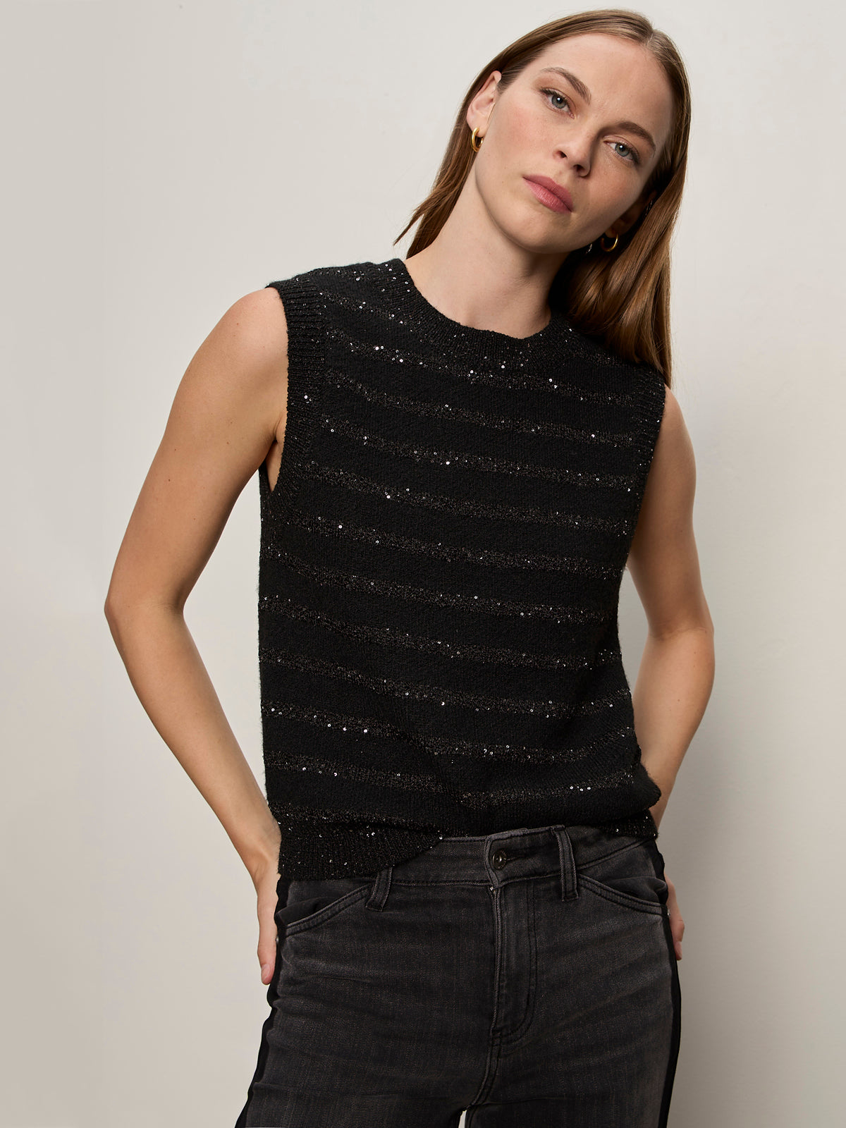 A woman with straight light brown hair models the Sanctuary Clothing shine sweater shell in black with black shine, paired with dark jeans. She stands against a plain light background, one hand in her pocket, looking slightly to the side.