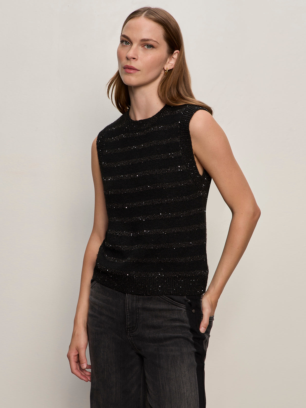 A woman with straight, light brown hair wears the Sanctuary Clothing shine sweater shell in black with black shine and dark jeans. She stands against a plain light background, looking to the side with a neutral expression.